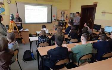 Chłopcy i dziewczyny słuchają nauczyciela, w tle urządzenia geodezyjne i prezentacja z hasłem "czym zajmuje się geodeta?"