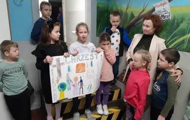 Uczniowie i uczennice klasy pierwszej i drugiej wraz z wychowawczynią prezentują plakat na temat chrztu.