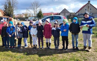 Zdjęcie przedstawia kolorowo ubraną grupę dzieci stojących na podw&oacute;rku szkolnym w p&oacute;lkolu uśmiechnięte dzieci ubrane na kolorowo. Jeden uczeń trzyma kartkę z zadaniami do rozwiązania.