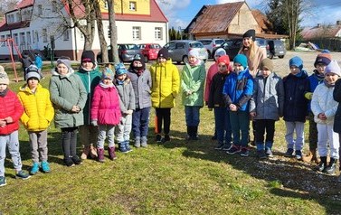 Zdjęcie przedstawia stojące na podw&oacute;rku szkolnym w p&oacute;lkolu uśmiechnięte dzieci ubrane na kolorowo. Za nimi stoi nauczycielka. W tle widać bawiące się na placu zabaw dzieci.