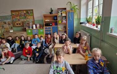 Dziewczynki i chłopcy siedzą w ławkach i na podłodze zasłuchani w wypowiedź nauczyciela. Mają  przypięte do ubrania ż&oacute;łte żonkile.