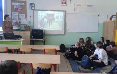 Uczniowie siedzą na podłodze w pomieszczeniu szkolnym. Na dużym ekranie oglądają film reklamujący ełcką szkołę średnią. Przy tablicy stoi nauczycielka.