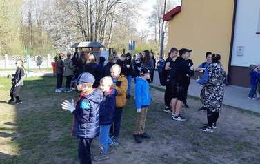Uczniowie szkoły przygotowują się do wyjścia w teren  w celu uprzątnięcia go. Na zdjęciu dzieci, młodzież i nauczyciele stojący w grupkach na boisku szkolnym. Trzymają worki na śmieci, zakładają ochronne rękawiczki.