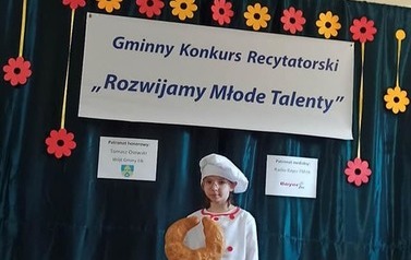 Dziewczynka w białym przebraniu piekarza, z ogromną czapą kucharską na głowie i rogalem w ręce recytuje wiersz.