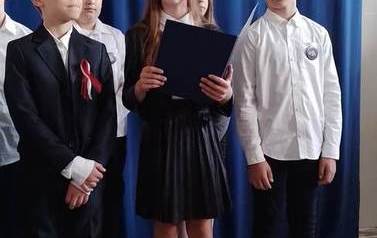 Dziewczynka trzyma w ręku granatową podkładkę, obok niej dwaj chłopcy. Wszyscy elegancko ubrani, stoją na środku sali.