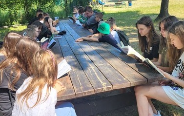 Uczniowie czytają książki, siedząc przy drewnianym stole na podw&oacute;rku szkolnym.