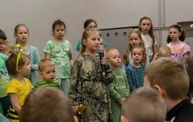 Dziewczynka trzyma mikrofon, obok dzieci przebrane za pszcz&oacute;łkę i konika polnego. Z tyłu grupa dzieci.