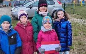 Czterech dziewczynki i jeden chłopiec stoją na podw&oacute;rku. Dziewczynka trzyma w rękach czerwoną kartkę.