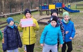 Czterech chłopc&oacute;w i dwie dziewczynki stoją na podw&oacute;rku. Dziewczynka trzyma w rękach r&oacute;żową kartkę.