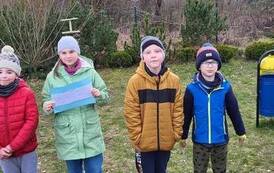 Trzech chłopc&oacute;w i jedna dziewczynka stoją na podw&oacute;rku. Dziewczynka trzyma w rękach niebieską kartkę.