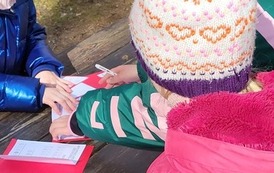 Zdjęcie przedstawia dziewczynki i chłopc&oacute;w zajętych rozwiązywaniem zadań zamieszczonych na kolorowych kartonikach. Dzieci zgromadzone wok&oacute;ł ławki w przyszkolnym parku.