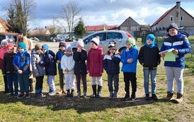 Zdjęcie przedstawia kolorowo ubraną grupę dzieci stojących na podw&oacute;rku szkolnym w p&oacute;lkolu uśmiechnięte dzieci ubrane na kolorowo. Jeden uczeń trzyma kartkę z zadaniami do rozwiązania.