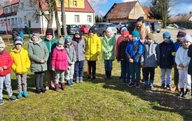 Zdjęcie przedstawia stojące na podw&oacute;rku szkolnym w p&oacute;lkolu uśmiechnięte dzieci ubrane na kolorowo. Za nimi stoi nauczycielka. W tle widać bawiące się na placu zabaw dzieci.