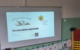 Na tablicy wyświetlony slajd prezentacji z hasłem "Bierzemy udział w akcji Żonkile". W tle nauczyciel wyświetlający i omawiający prezentację.