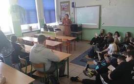 Zdjęcie przedstawia grupę uczni&oacute;w siedzących w ławkach i na podłodze w sali szkolnej. Oglądają ulotki, słuchają wypowiedzi nauczycielki.