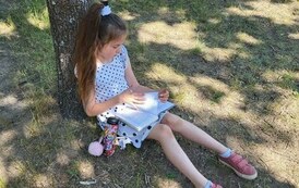 Pod drzewem siedzi dziewczynka z książką na kolanach.