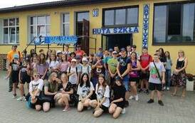 Zdjęcie grupowe uczestnik&oacute;w wycieczki stojących przed budynkiem Huty Szkła Artystycznego w Olsztynku.